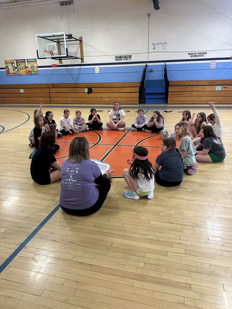 Group of elementary-aged girls and coaches sitting in a circle on a gym floor, raising hands and participating in a group discussion during Girls on the Run practice.