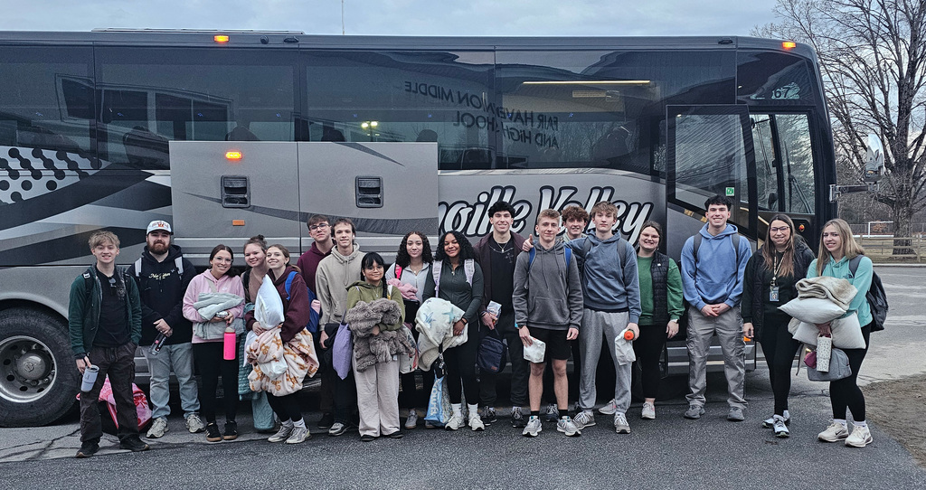 Group of high school students standing together in a parking lot in front of a coach bus, holding bags and blankets as they prepare to depart for a school trip.