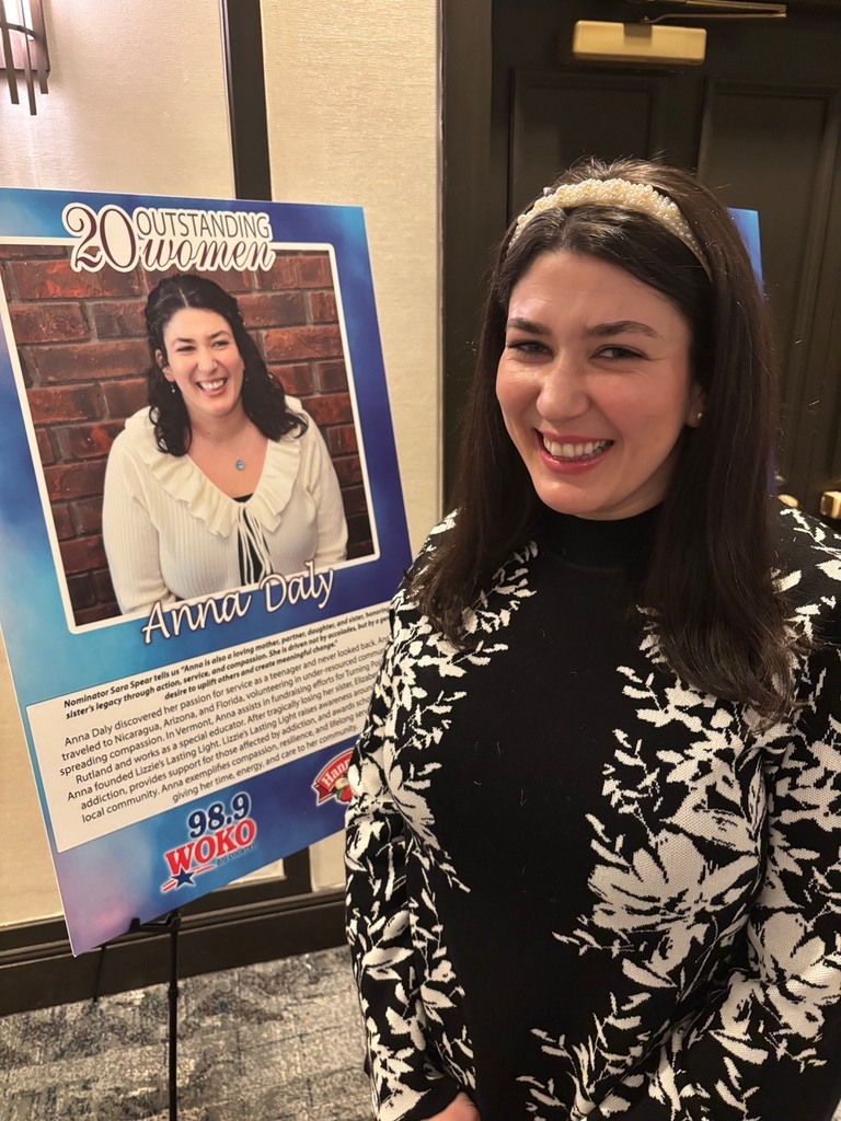 A woman stands smiling beside a display poster that reads “20 Outstanding Women” featuring her photo and name, Anna Daly.