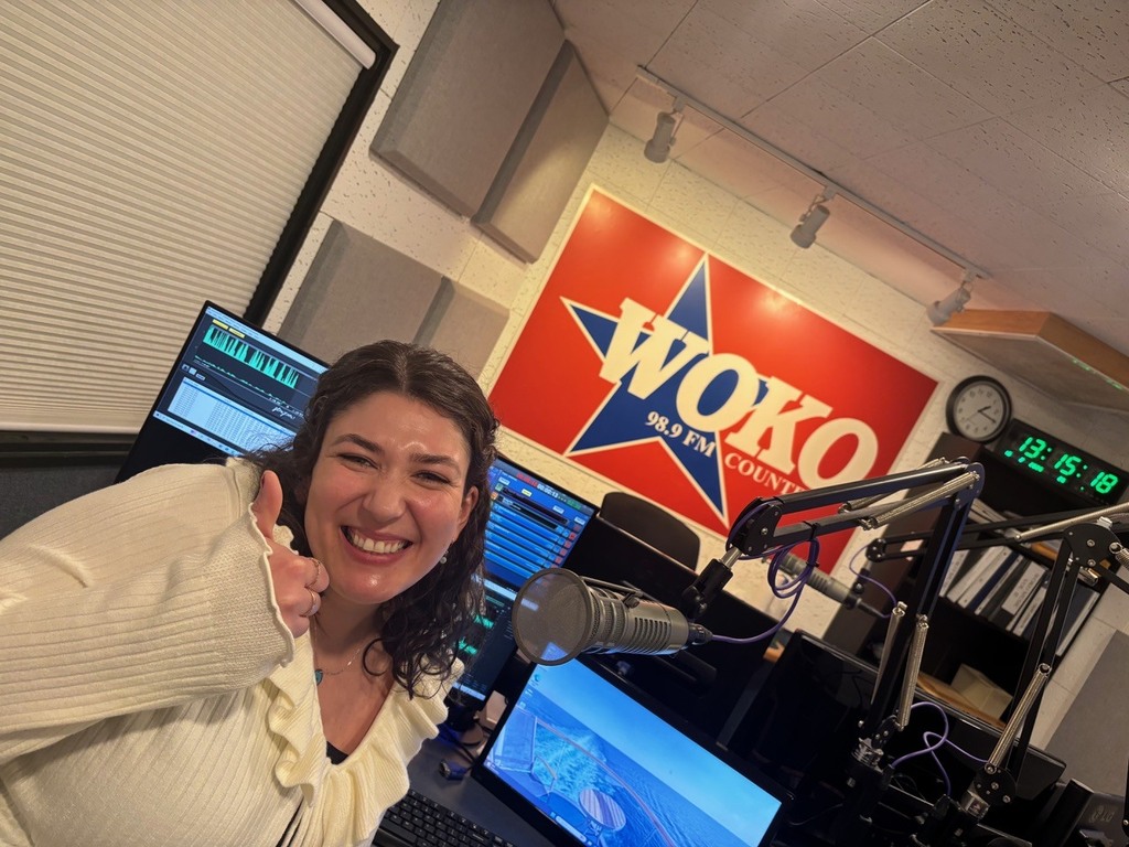 A smiling woman gives a thumbs-up while sitting in a radio studio in front of a microphone, with a “WOKO 98.9 FM” sign and broadcast equipment visible behind her.