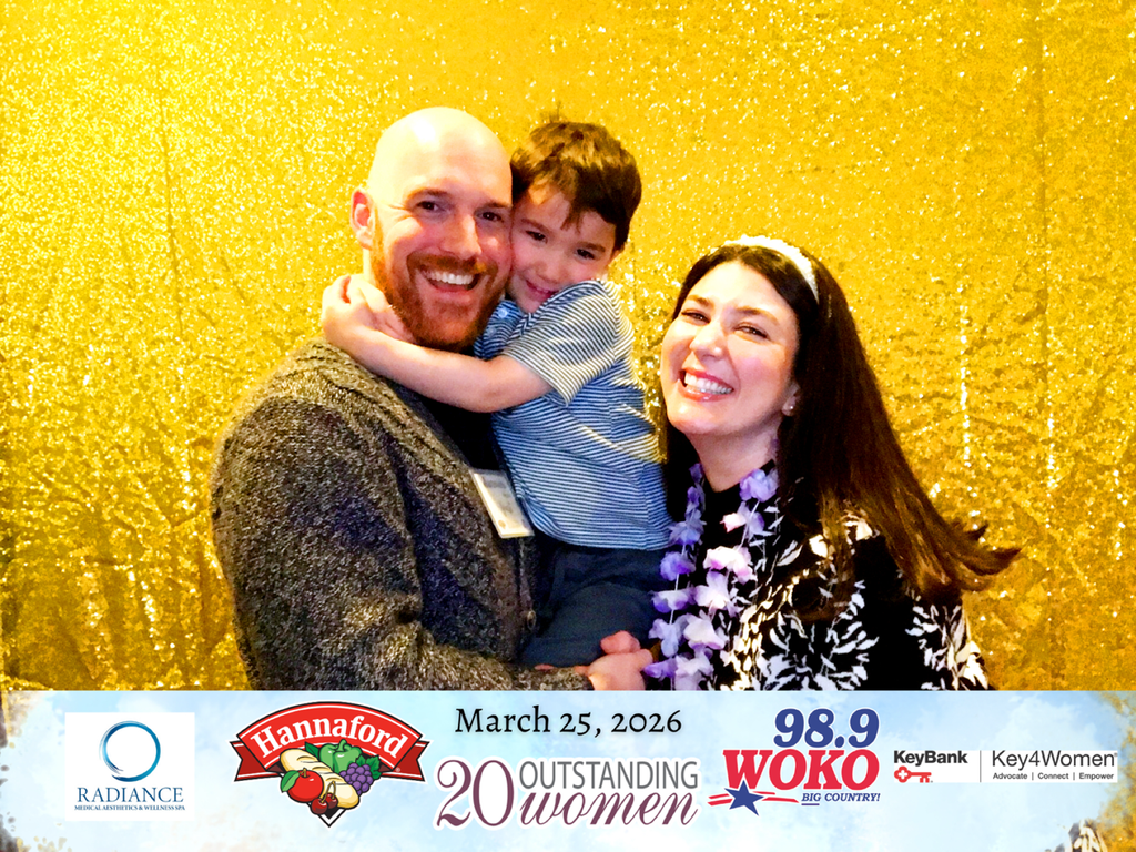 A woman stands with a man and a young child in front of a gold backdrop at an awards event, all smiling as they pose together.