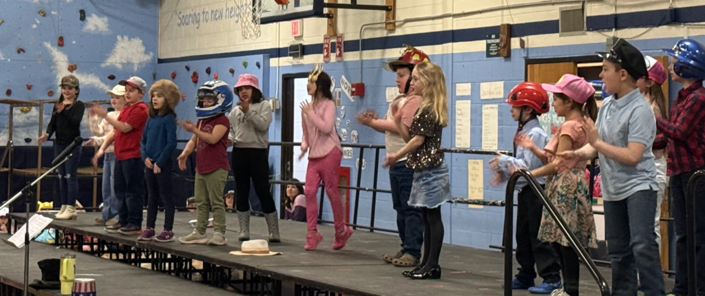 Elementary students perform on stage wearing a variety of hats and costumes, clapping and singing during a musical.