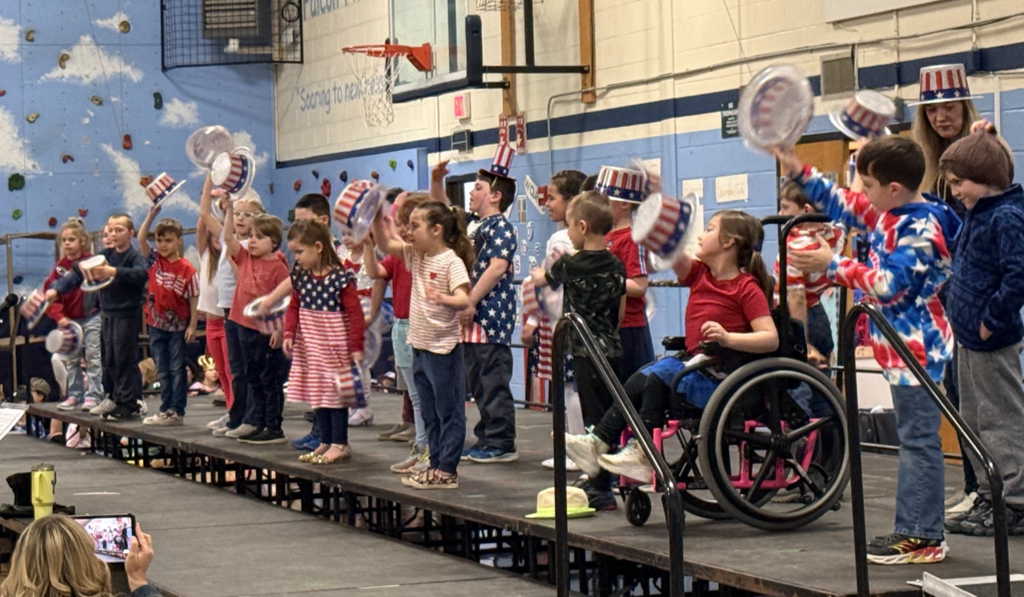 Students on stage wave patriotic-themed hats while performing, with one student in a wheelchair included in the group.