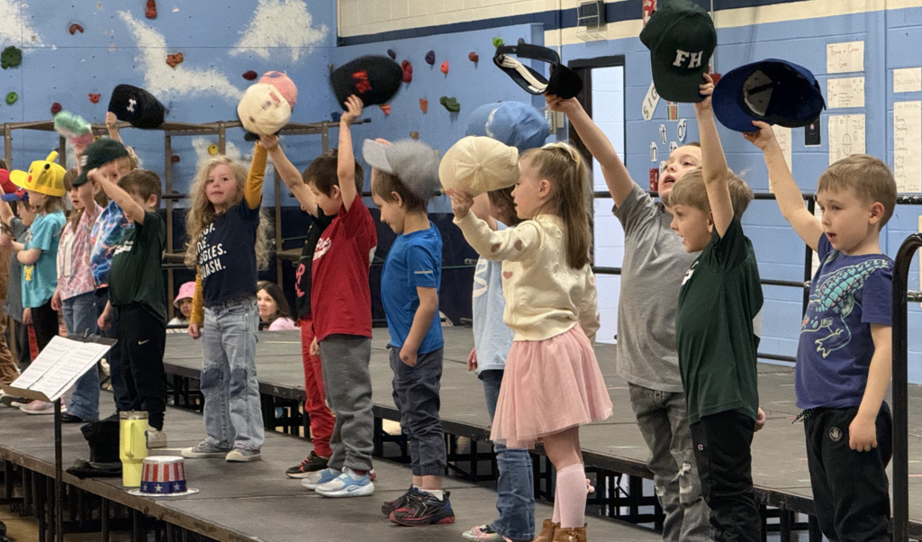 Children stand in a row on stage holding up different hats as part of a musical performance in a school gym.