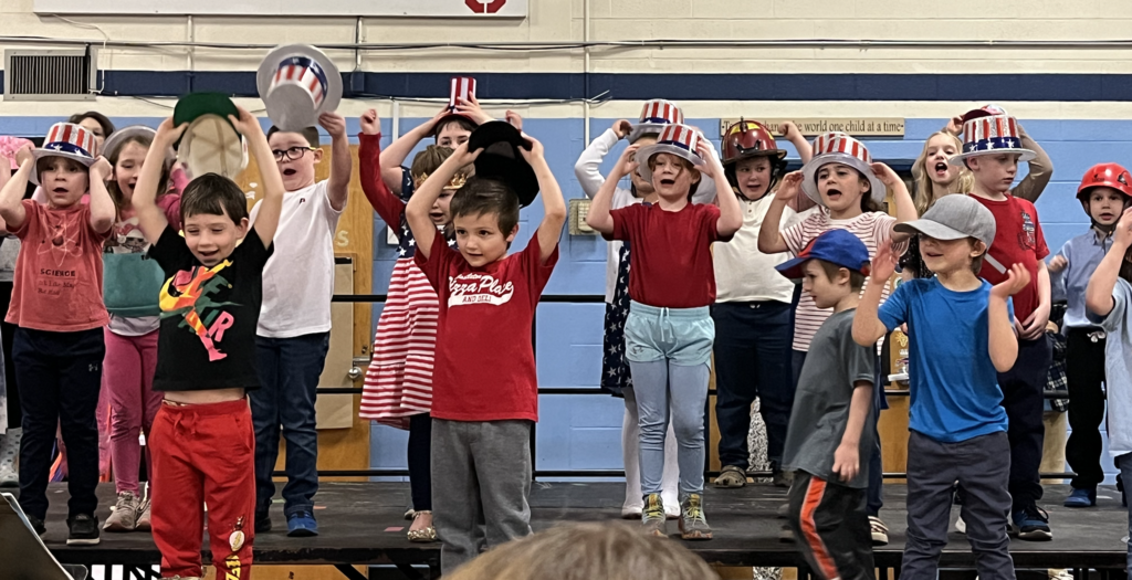 Young students on a stage wearing red, white, and blue hats, singing and raising their hats during a school performance in a gym.
