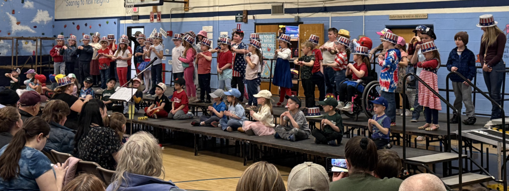 A large group of young students perform on stage in front of an audience seated in a school gymnasium.