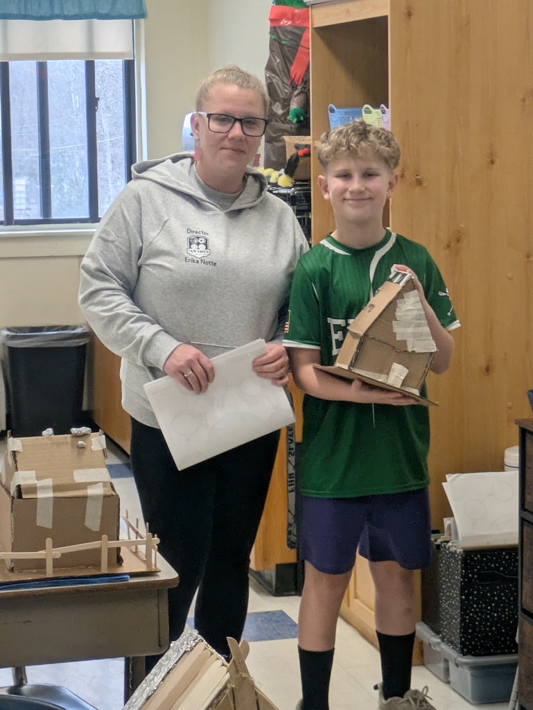 A vertical photograph shows a woman and a young boy standing together in a classroom, posing with school projects made of cardboard.