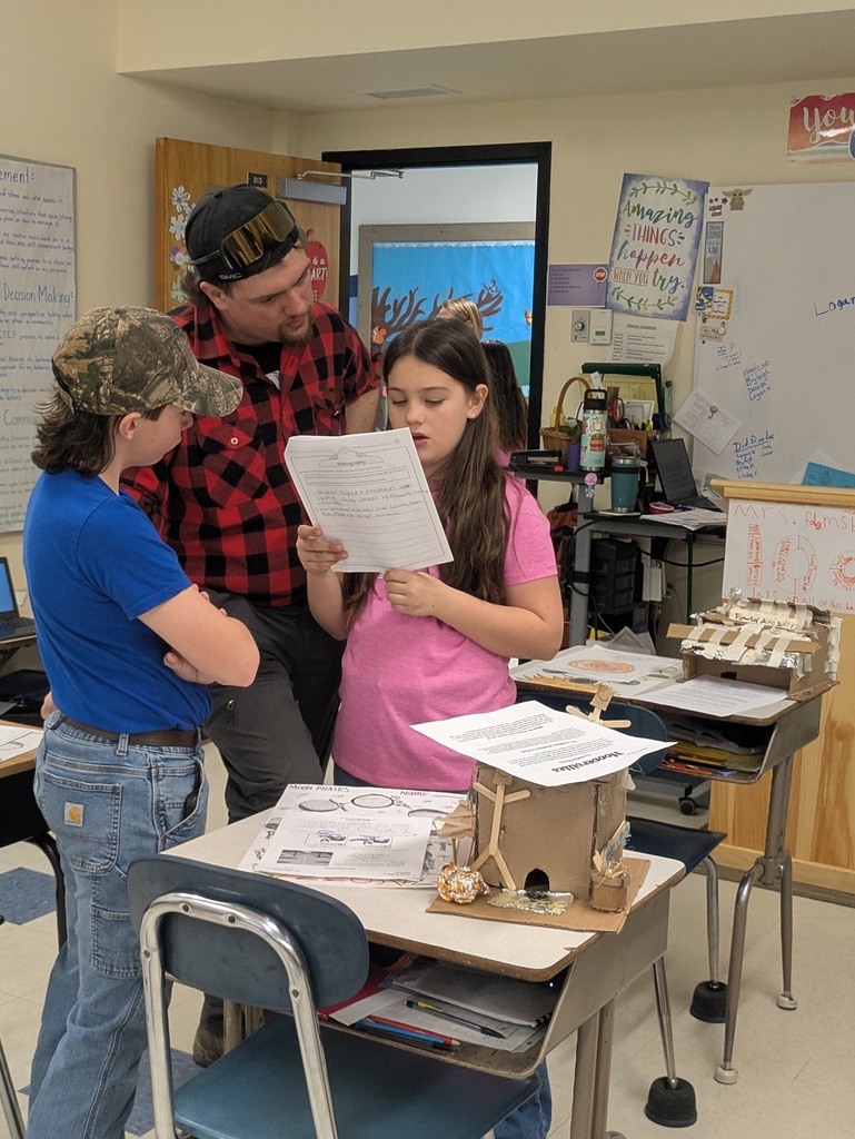 Gemini said Image Summary A vertical photograph shows a man and two children reviewing schoolwork in a classroom. The girl in the center appears to be presenting or reading from a worksheet while the man and another child listen attentively.