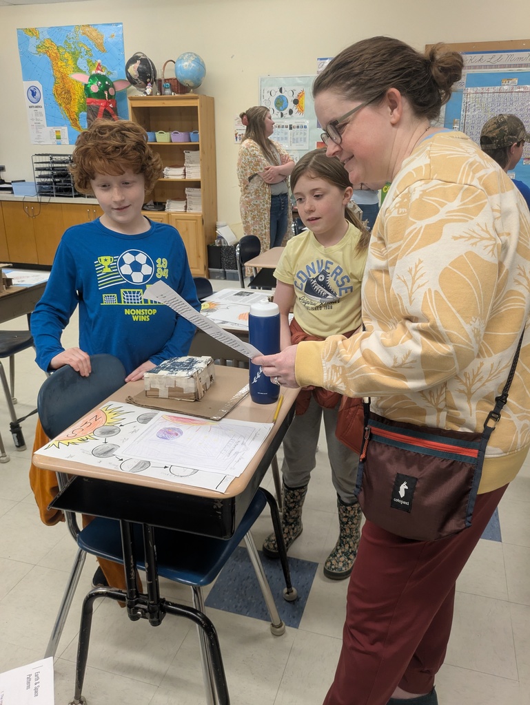 A vertical indoor photograph shows a woman and two children in a classroom, looking at school projects displayed on a student desk.