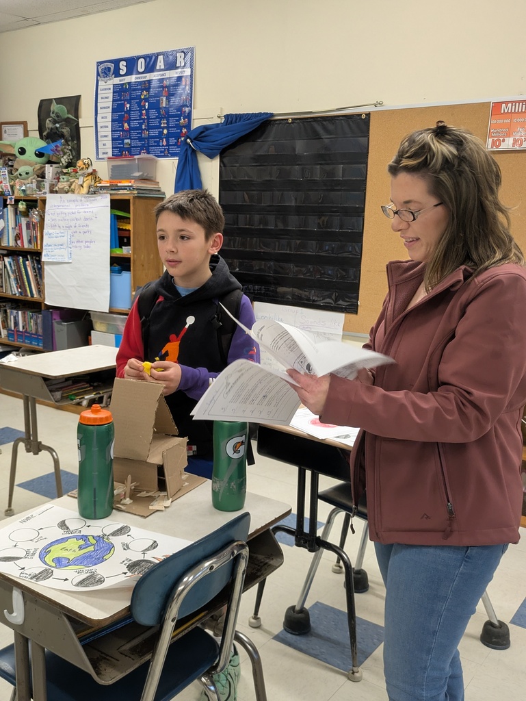 A vertical photograph shows a woman in a classroom flipping through a student’s packet while a young boy stands beside his project display.