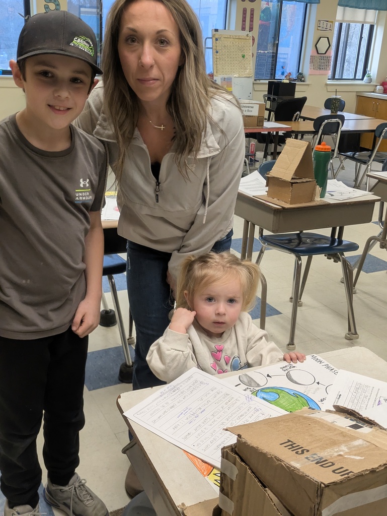 A vertical indoor photograph captures a woman and two children in a primary school classroom, likely during a project presentation or open house.