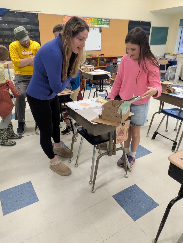 A vertical photograph shows a woman and a young girl interacting over a school project in a classroom. The woman leans in with an enthusiastic expression as the girl displays her work.
