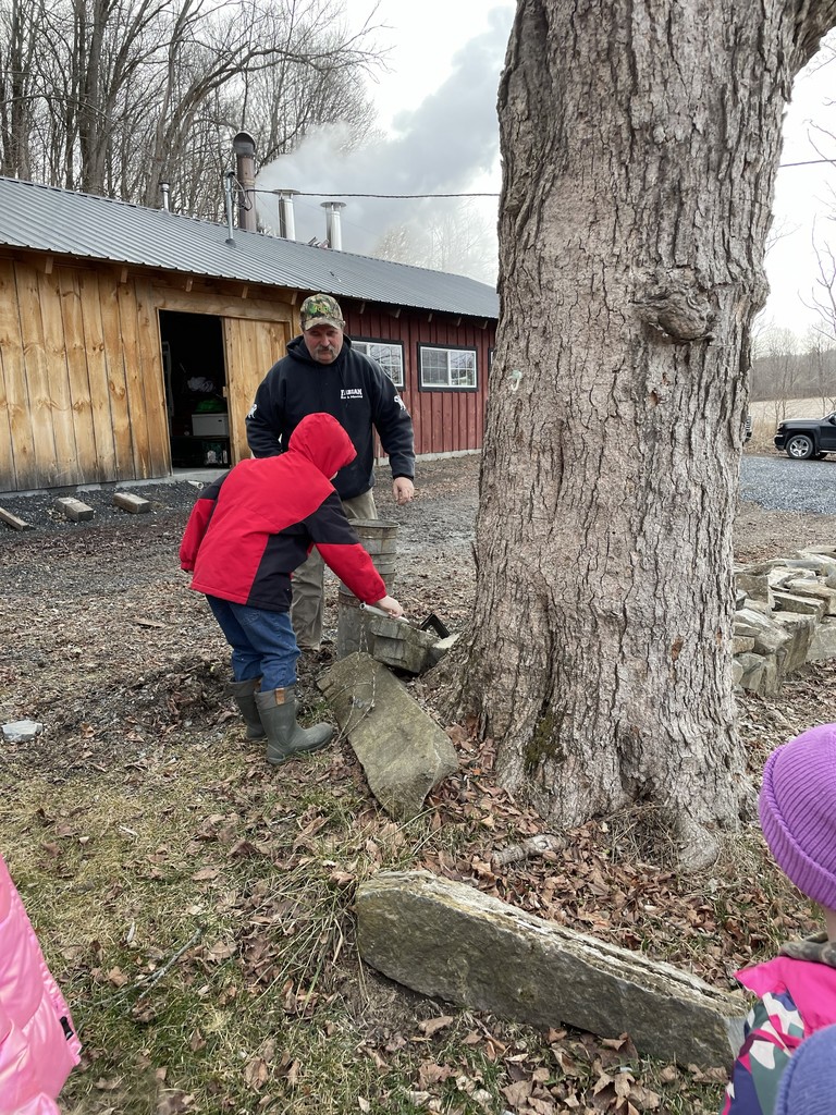 A high-angle full shot shows two men standing on dry leaves in front of a red wooden shed next to a very tall tree, and they appear to be pouring water. The man in front wears a red jacket, blue pants, and green boots, and he is holding a container over a puddle of water with a wire near the tree. The second man is standing right behind him wearing a green hat, a black shirt, and beige pants and has a light beard and mustache. His mouth is closed, but he appears to be talking or looking at the water. The tree has dark brown bark, and the shed is red with small windows and a chimney, with grey steam coming from it. The leaves are light to dark brown and cover the ground and rocks. The man in front is looking down at his bucket, while the second man looks forward at the tree with a concentrated expression. The camera is steady.