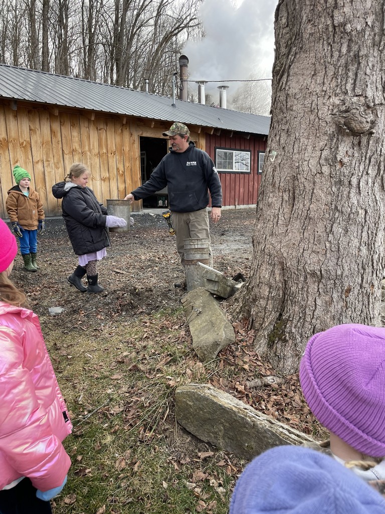 A candid, medium shot photo of a man and two children tending to maple sap buckets on a large tree during a late winter/early spring day. The man, wearing a camouflage baseball cap, black sweatshirt with a white dog logo, and beige cargo pants, stands on the right, holding a metal rod. The first child, facing away from the camera on the left, wears a vibrant orange beanie, a red and black plaid flannel jacket, and camouflage pants, looking at a sap bucket. The second child, in the center, wears a purple knit hat and a bright pink and purple patterned coat, and is looking at a metal bucket near her. Behind them is a large, rough-barked tree, and beyond that, a rustic red-sided building with a metal roof is partially obscured by trees. Steam rises from a chimney on the building, indicating sap boiling. There is a metal ATV parked near the building. The ground is dry grass and fallen leaves with stacked firewood nearby. The lighting is diffused under an overcast sky.