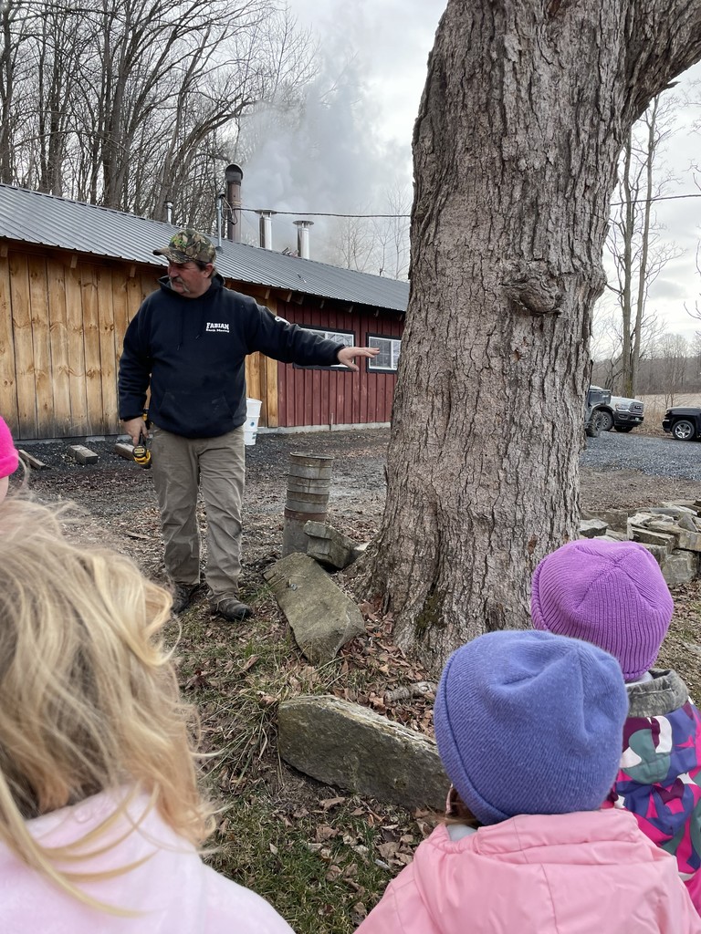 A man in a camouflage hat and black hoodie stands outdoors, gesturing toward a large, mature tree with rough bark. In the foreground, the backs of three children wearing colorful winter hats (pink, purple, and blue) are visible as they watch him. In the background, a rustic red wooden building with a metal roof emits white smoke from its chimney.