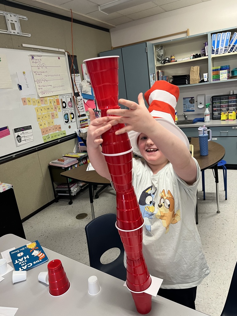 A smiling boy wearing a Bluey character shirt and a tall, red and white striped "The Cat in the Hat" style top hat with a white brim, uses both hands to stabilize a towering stack of red solo cups. The top red cup rests on a white index card. In the foreground, a gray table holds several red and white cups and a children's book titled "The Cat in the Hat." The background features a classroom setting with a whiteboard displaying math problems, and a shelf with plastic bins