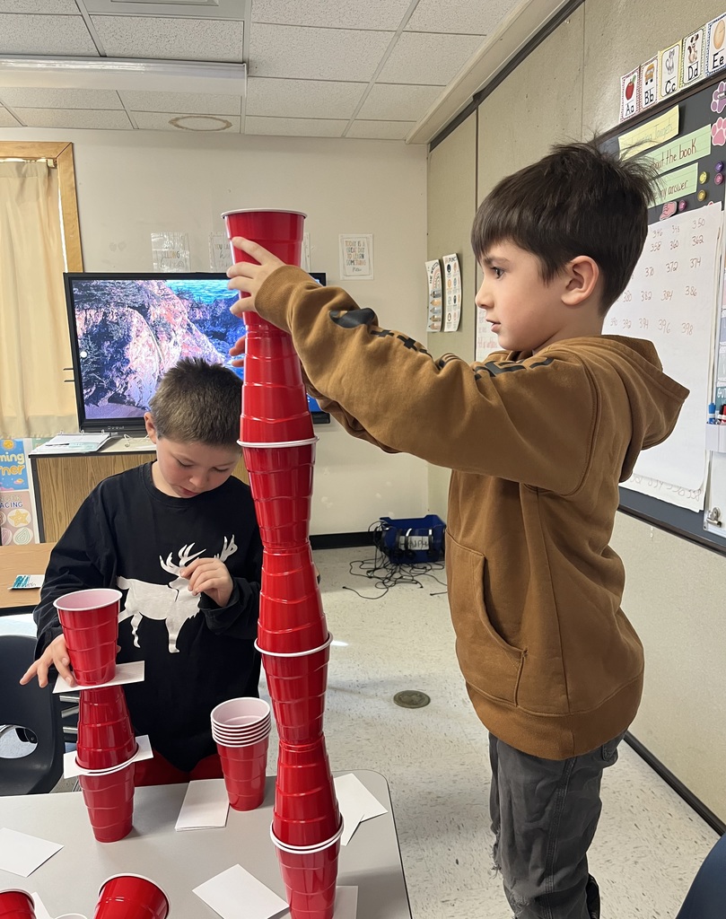 : A young boy in a brown hoodie carefully places a red cup onto a tall stack, using both hands to steady the structure. Another student in a black shirt watches closely from the side. 