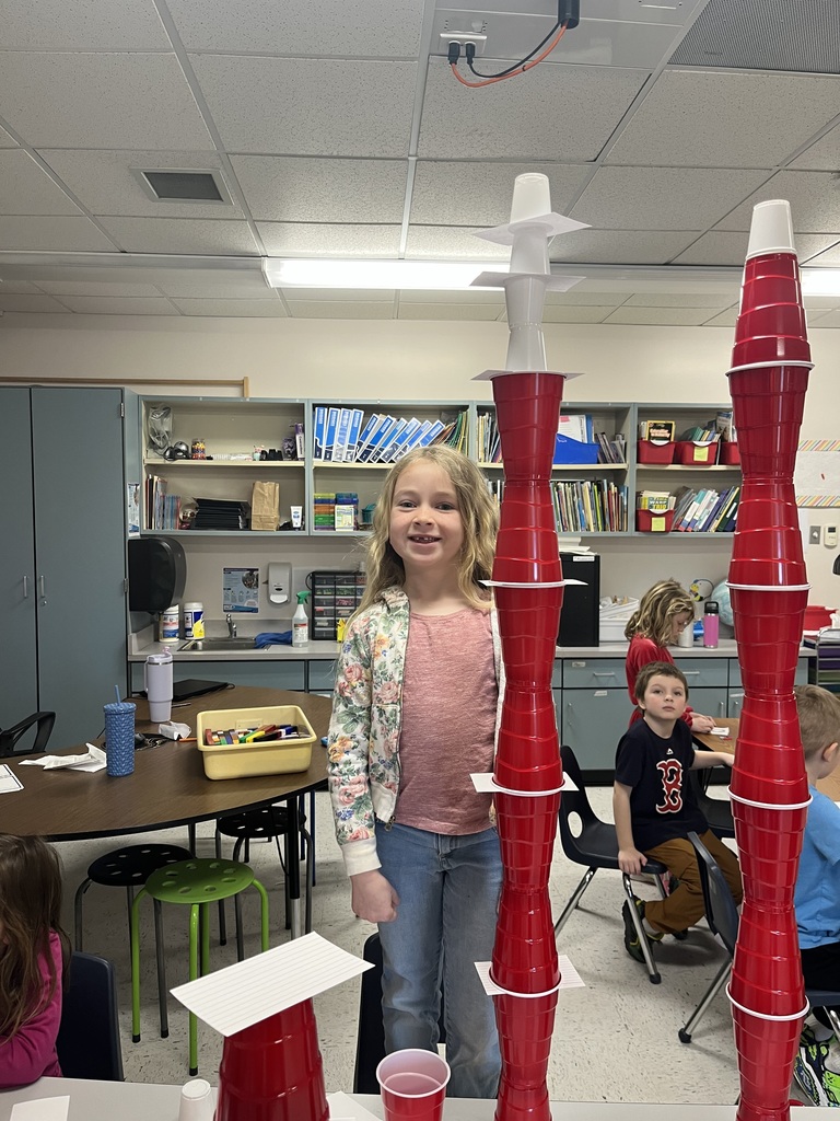 A smiling girl stands between two impressively tall towers made of red cups and index cards. The tower on the left is topped with smaller white cups, nearly reaching the height of the classroom bookshelves.