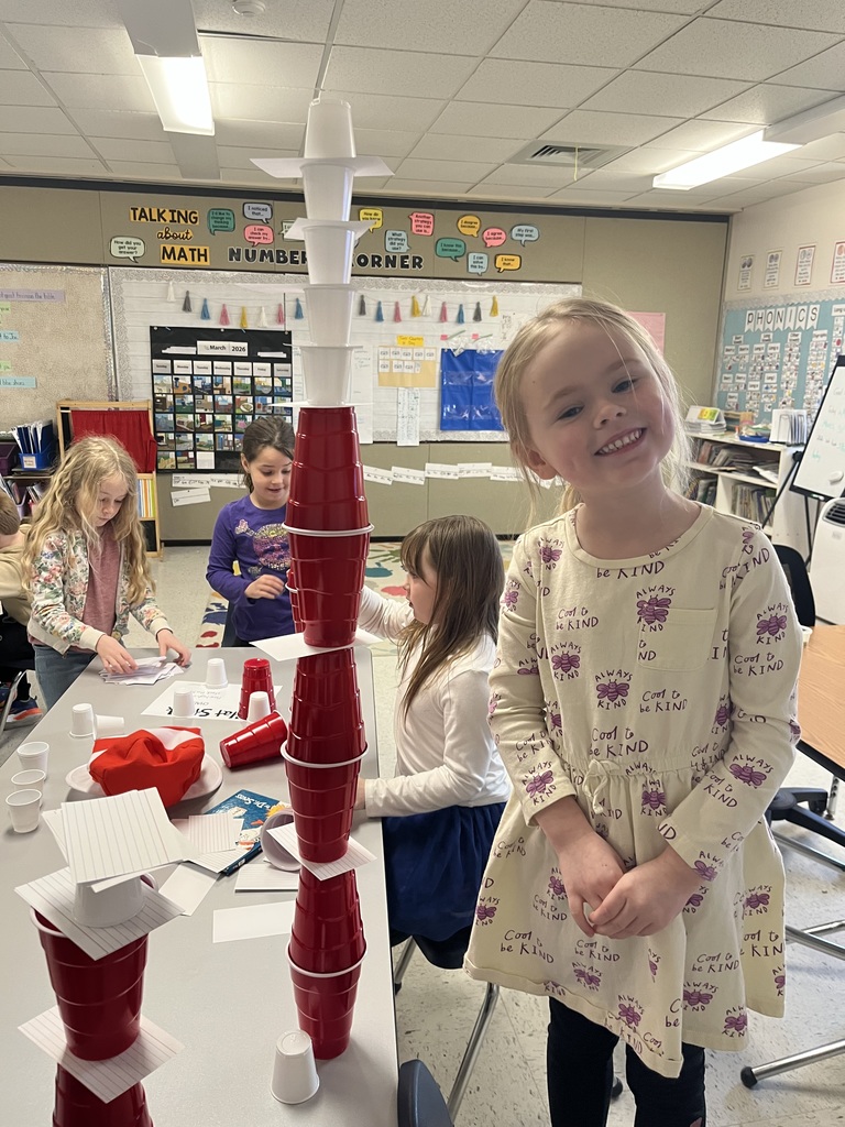 : A young girl in a "Cool to be Kind" dress smiles next to a tall, narrow tower of red cups that transitions into white cups at the top. Other students are visible in the background working on their own structures.  