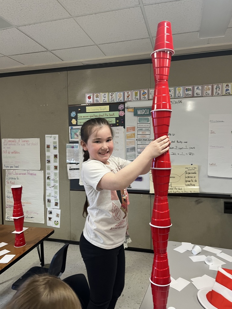 A smiling girl stands between two impressively tall towers made of red cups and index cards. The tower on the left is topped with smaller white cups, nearly reaching the height of the classroom bookshelves.  