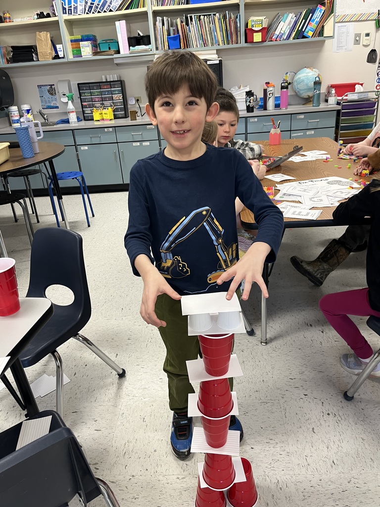A young boy in a navy long-sleeve shirt with an excavator graphic stands behind a carefully balanced tower of red plastic cups and white index cards. He has his hands poised near the top, concentrating on his engineering feat. 