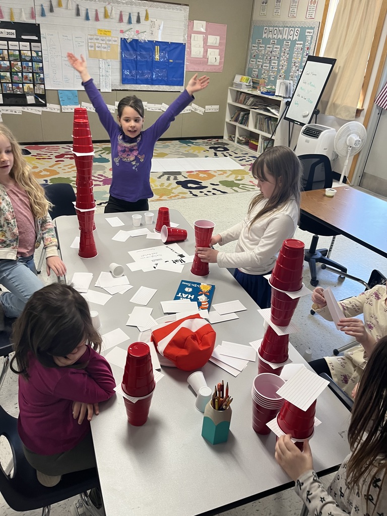 A group of children collaborates at a table covered in red cups, white cups, and index cards. One girl in a purple shirt has her arms raised in celebration next to a tall red cup tower, while a copy of The Cat in the Hat and a red-and-white striped hat sit on the table.  