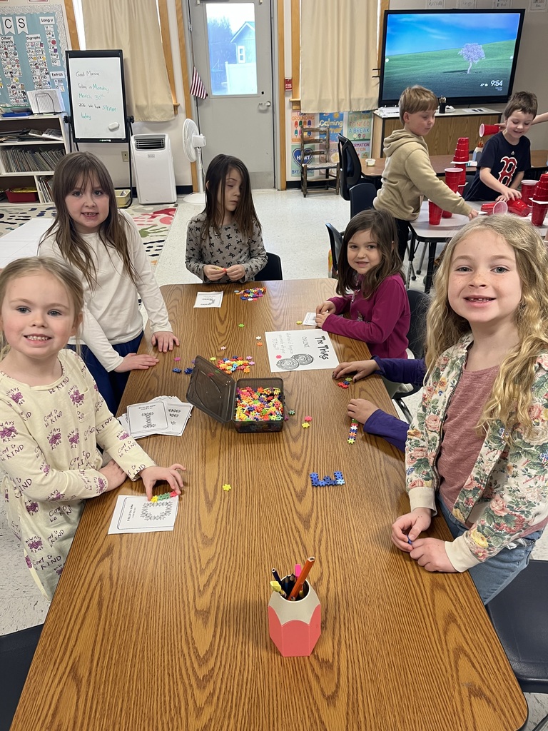  Two smiling girls stand in the foreground of a busy classroom. Behind them, other students are focused on stacking red and white plastic cups into tall towers on a gray table.  IMG_4972.jpg: A group of children collaborates at a table covered in red cups, white cups, and index cards. One girl in a purple shirt has her arms raised in celebration next to a tall red cup tower, while a copy of The Cat in the Hat and a red-and-white striped hat sit on the table. 