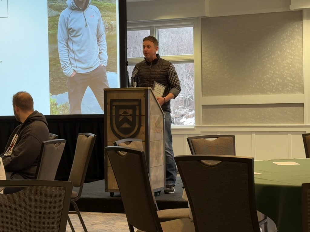 A man stands at a podium holding an award and speaking into a microphone during a conference event, with a projection screen behind him showing his recognition.