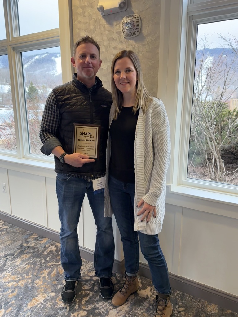 A man holds a plaque labeled “SHAPE Vermont Physical Education Teacher of the Year 2026” while standing next to a woman indoors near large windows, both smiling for the camera.
