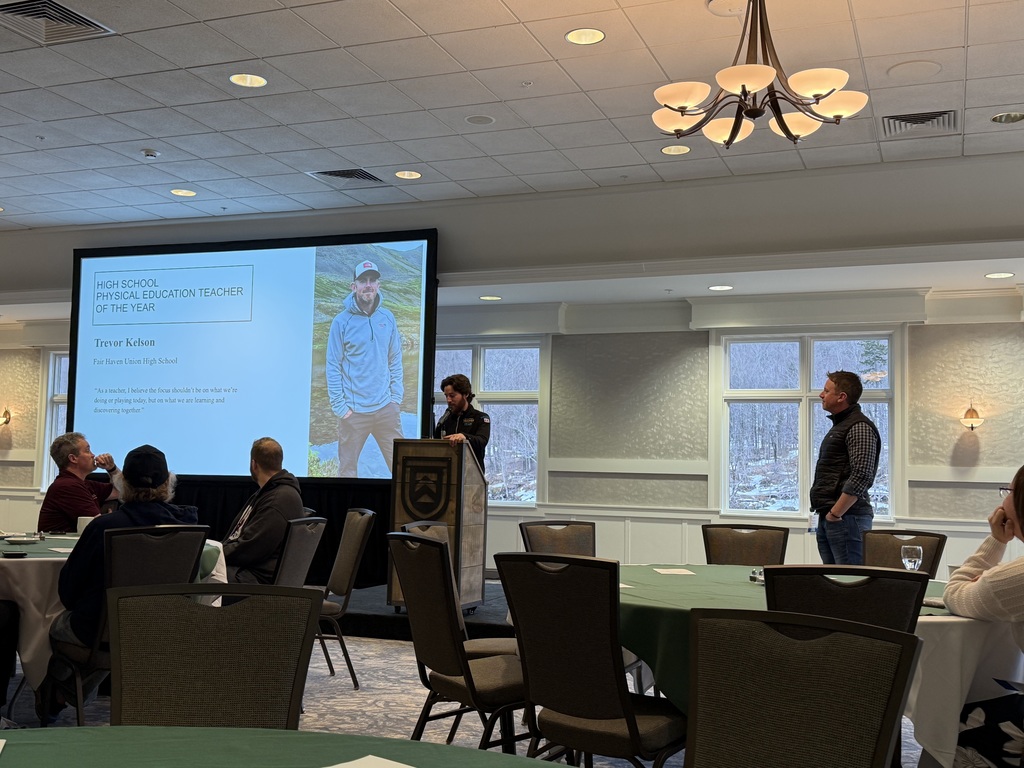 A man stands at a podium speaking during an awards event in a conference room while a large screen behind him displays “High School Physical Education Teacher of the Year” with his name and photo. Attendees sit at round tables listening.