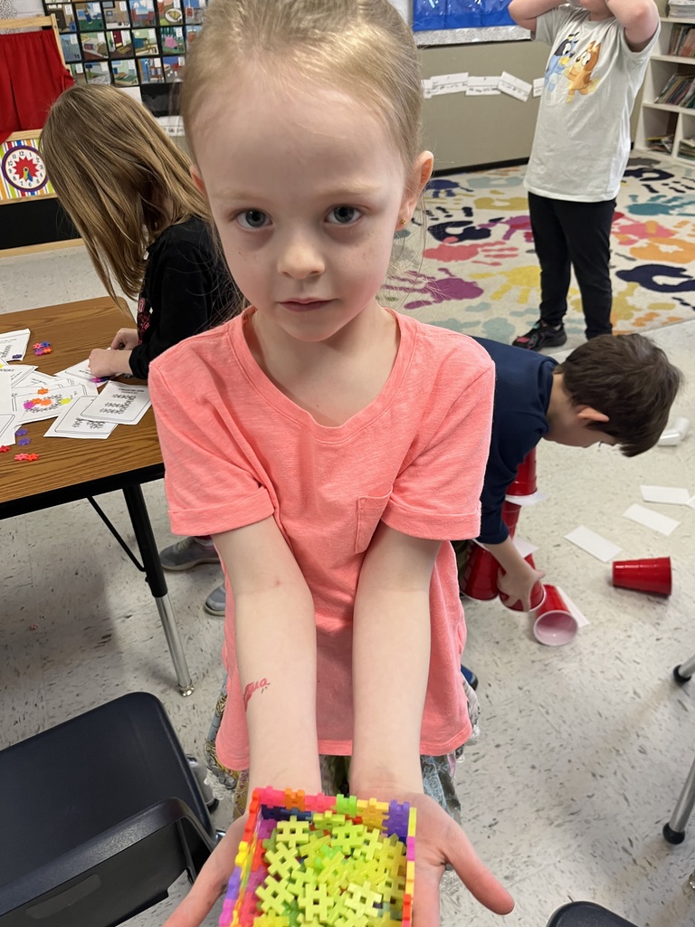  A young girl with light hair is looking intensely at the camera, wearing a solid pink t-shirt and holding out two cupped hands filled with a small handful of colorful yellow and green plus-sign connecting blocks. Behind her, a busy classroom shows other students engaged in activities and a multi-colored rug. 