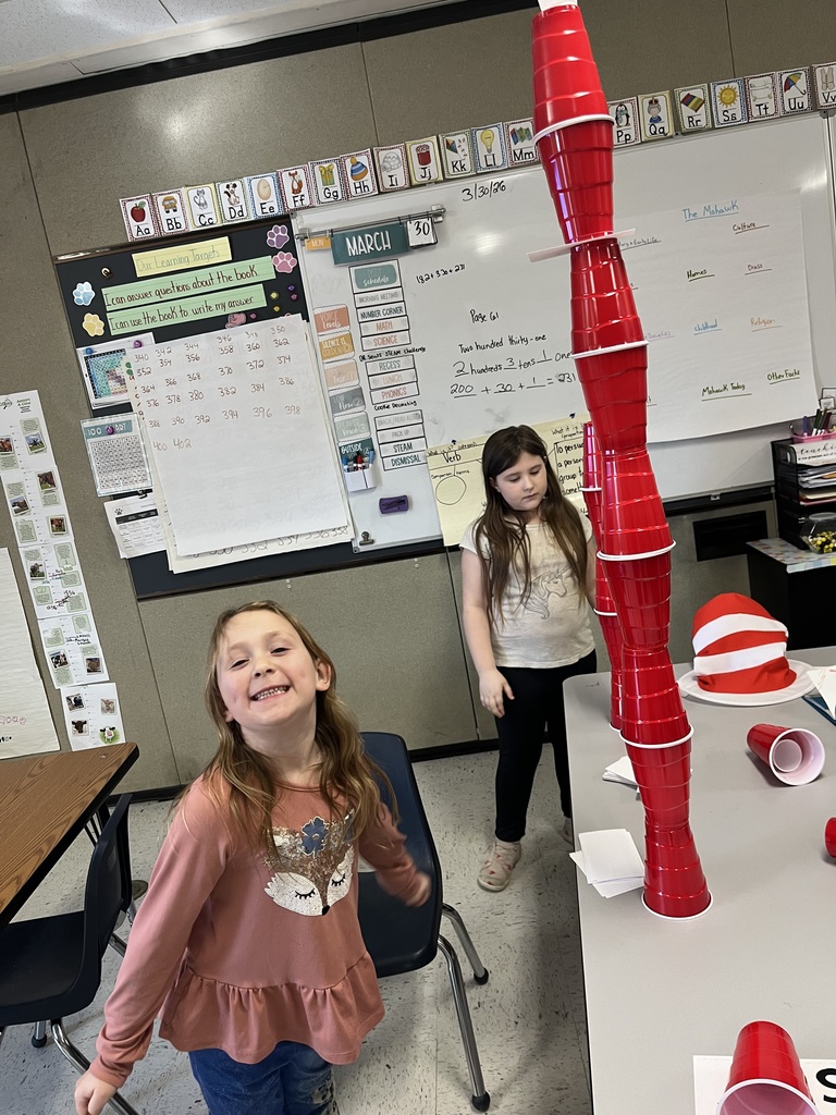  A high-angle view captures a girl in a pink ruffled fox sweater grinning broadly and looking straight up at the camera with a cheerful, joyful expression in a classroom setting. To the left, a tall tower of red and white cups is balanced, reaching high above a desk, while a large whiteboard with lesson notes and a "March" date covers the background.  