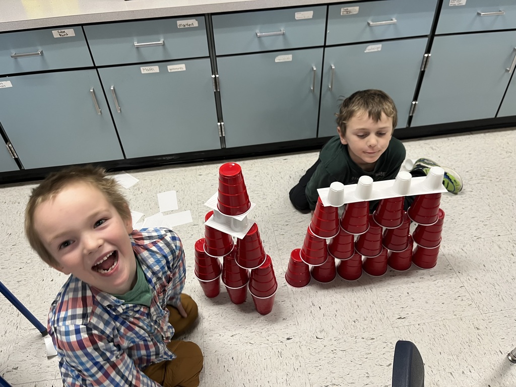 A smiling boy in a plaid shirt is sitting on the floor, looking directly at the camera with an open-mouthed expression of excitement next to a tower of stacked red cups. A second child is seated slightly further back, focused on building an adjacent structure with white and red cups, while labeled classroom cabinets fill the background. 