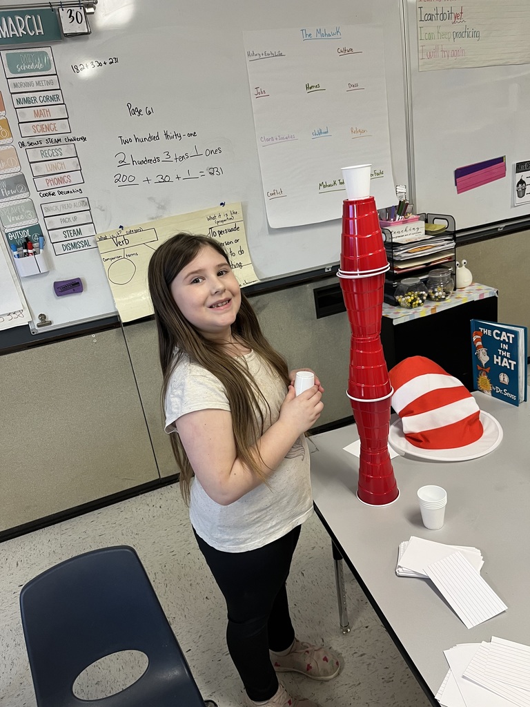 A young girl with long, light brown hair stands near a classroom desk, smiling broadly as she holds two stacked white paper cups. She is wearing a white graphic t-shirt and black pants, and on the desk next to her is a tall tower built of stacked red and white cups, a plush red-and-white hat, and a book titled "The Cat in the Hat." A whiteboard with lesson notes forms the background.