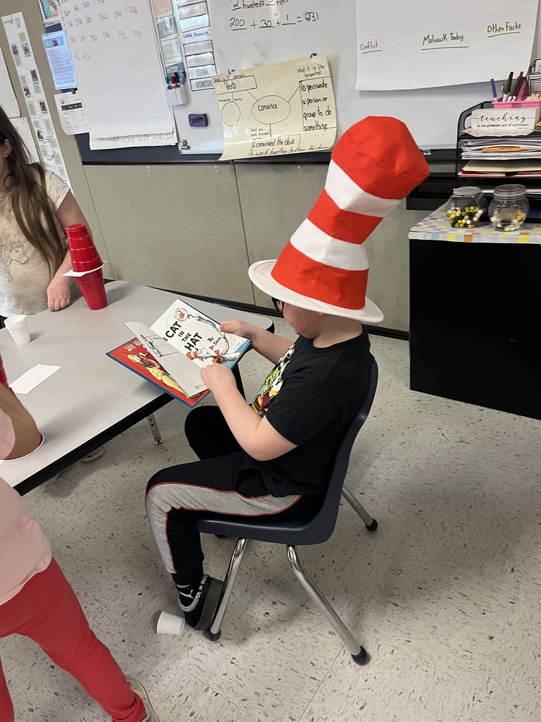 A child is seated at a school table, seen from a high side angle, engrossed in reading a worn hardcover copy of "The Cat in the Hat." He is wearing a tall, iconic red-and-white striped hat and black pants with gray trim, while other classroom details are visible around him. 