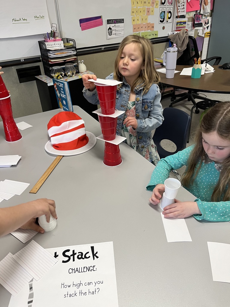  A group of children at a classroom table are participating in a "Hat Stack Challenge," with a sign on the table posing the question, "How high can you stack the hat?" A girl in a denim jacket carefully stacks red cups separated by index cards. Another girl nearby is attempting a smaller stack of white paper cups. A tall, red-and-white striped plush hat rests on the table as inspiration. 
