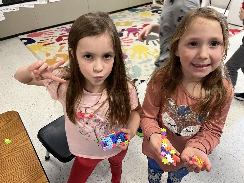  A top-down view of two smiling young girls in a classroom. The girl on the left is making a peace sign, wearing a pink shirt with a llama graphic, and holding a handful of colorful connecting blocks. The girl on the right, wearing a peach-colored graphic shirt, shows off a construction built with similar blocks. A colorful rug with handprints is visible in the background.  