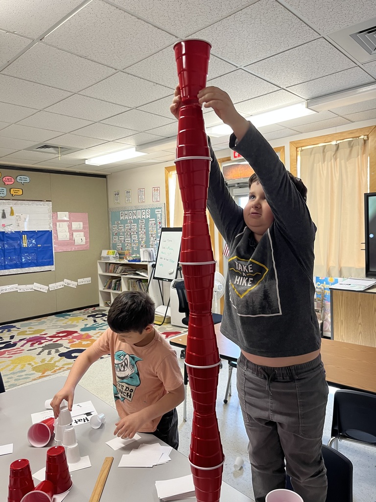  Two boys are working with disposable cups at a classroom table. The boy in the foreground, wearing a grey t-shirt that says "TAKE A HIKE," is reaching up high to add a red plastic cup to the top of a exceptionally tall tower made of multiple red and white cups. The second boy stands focused nearby, working with a separate collection of white cups.