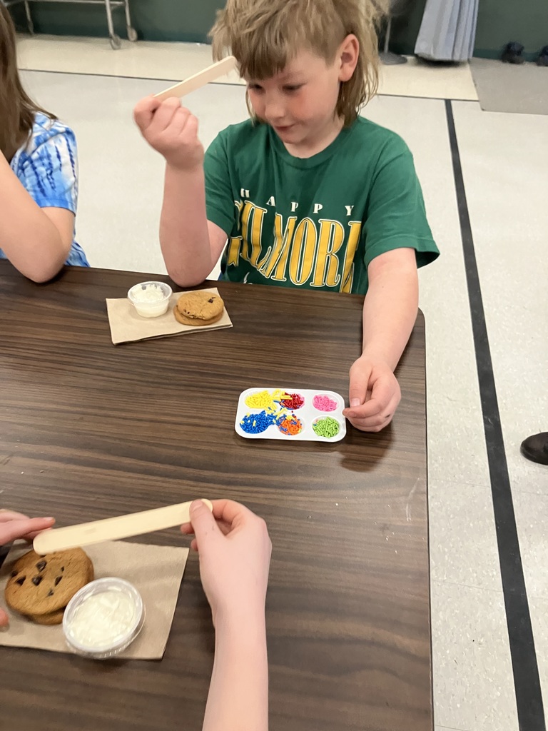 A child wearing a green "Happy Gilmore" t-shirt sits at a wooden table, holding a wooden craft stick. In front of them are two chocolate chip cookies on a napkin, a small container of white frosting, and a white palette filled with colorful sprinkles in various shades like blue, orange, and pink.  IMG_3155.jpg: Two young boys sit at a long cafeteria table prepared for cookie decorating. Each has a set of chocolate chip cookies, a cup of frosting, and a wooden spreader. A shared palette of bright sprinkles sits between them on the dark wood surface. 