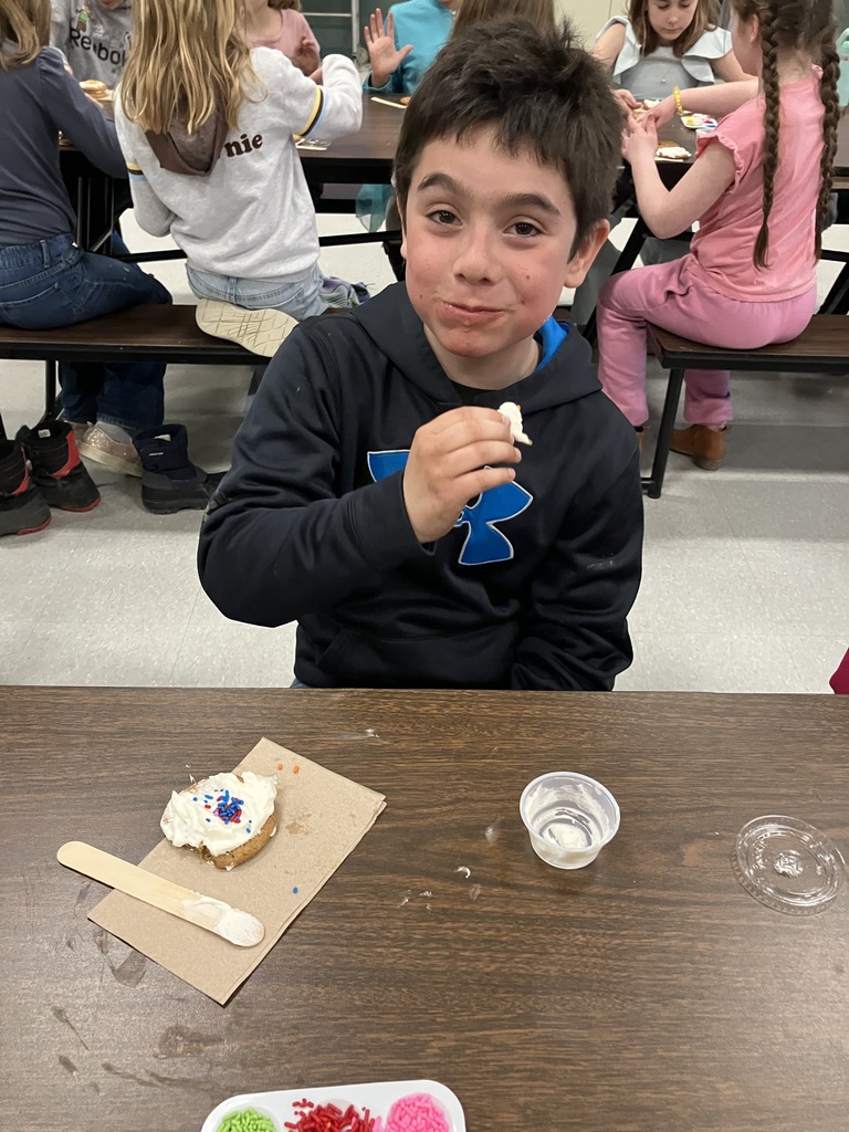 A boy in a black hoodie takes a bite of a cookie he just decorated. On the napkin in front of him is a chocolate chip cookie covered in thick white frosting and topped with red and blue sprinkles. 