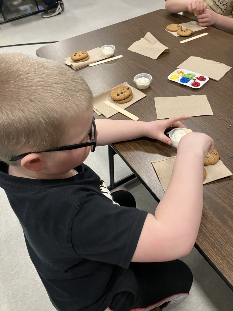 A high-angle shot showing a young boy with glasses beginning to decorate his cookies. Several other cookie-decorating stations are visible along the table, each equipped with napkins, cookies, frosting, and wooden sticks. 