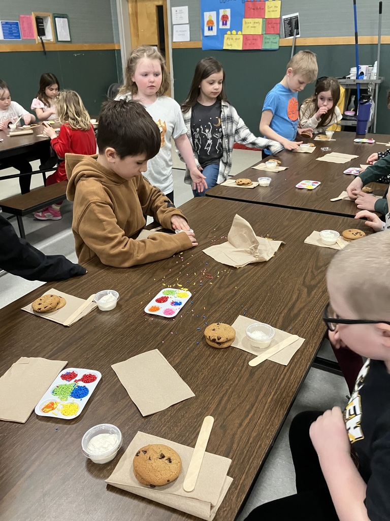 : A high-angle shot showing a young boy with glasses beginning to decorate his cookies. Several other cookie-decorating stations are visible along the table, each equipped with napkins, cookies, frosting, and wooden sticks. 