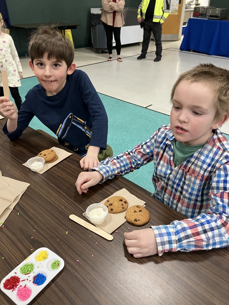 : Two young boys sit at a long cafeteria table prepared for cookie decorating. Each has a set of chocolate chip cookies, a cup of frosting, and a wooden spreader. A shared palette of bright sprinkles sits between them on the dark wood surface. 