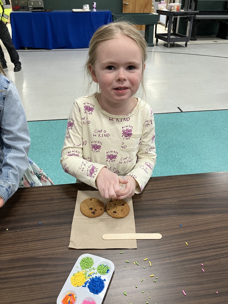  A young girl in a cream-colored "Be Kind" patterned shirt smiles at the camera. She has two chocolate chip cookies and a wooden stick ready on a napkin, with a palette of vibrant sprinkles nearby.  