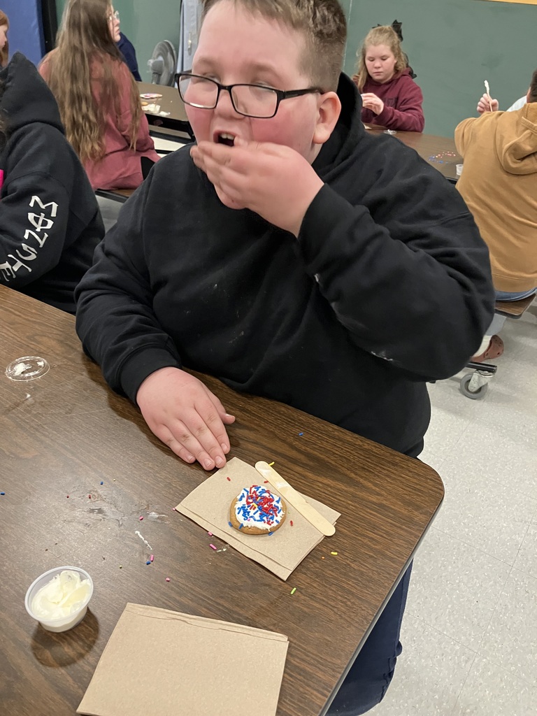 A boy in a black hoodie takes a bite of a cookie he just decorated. On the napkin in front of him is a chocolate chip cookie covered in thick white frosting and topped with red and blue sprinkles. 