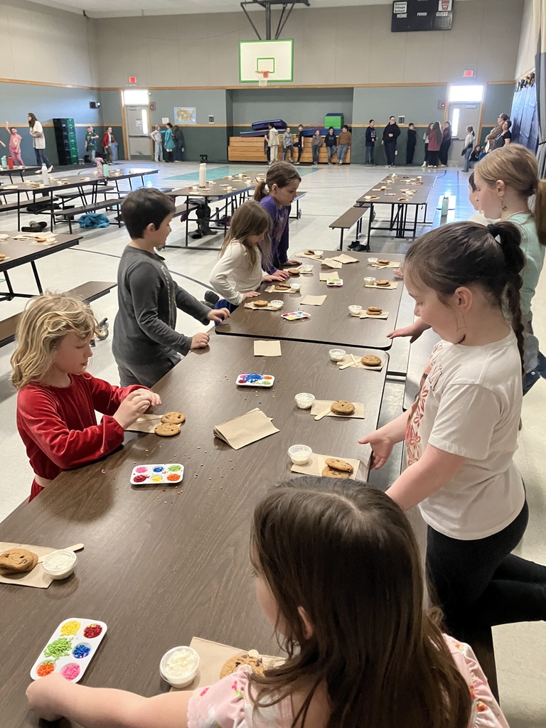 A wide shot of a school gymnasium or cafeteria where several elementary-aged children are standing at long brown tables. Each child has a workstation featuring two chocolate chip cookies on a napkin, a small cup of white frosting, a wooden spreader, and a plastic palette filled with various colorful sprinkles. In the background, other students and adults are visible near the gym walls. 