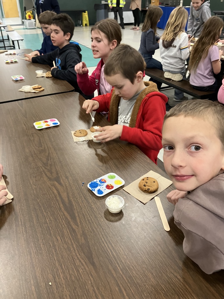  A wide shot of a school gymnasium or cafeteria where several elementary-aged children are standing at long brown tables. Each child has a workstation featuring two chocolate chip cookies on a napkin, a small cup of white frosting, a wooden spreader, and a plastic palette filled with various colorful sprinkles. In the background, other students and adults are visible near the gym walls.  