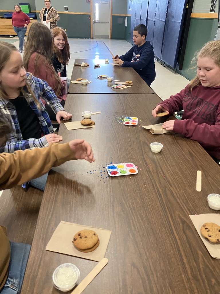 Children sit along a long table in a gymnasium, beginning to decorate their cookies. A girl in a blue plaid shirt looks toward the center of the table while a boy in a navy hoodie focuses on his cookies in the background. A palette of colorful sprinkles is shared between students, and several small cups of frosting and wooden sticks are scattered across the table.  