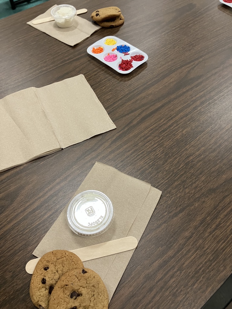  A high-angle, close-up shot of a dark wood-grain table prepared for a cookie decorating activity. Two workstations are visible; each includes two chocolate chip cookies, a brown paper napkin, a small plastic lidded cup of white frosting, and a wooden craft stick. A white plastic palette sits between the stations, containing six compartments filled with vibrant sprinkles in yellow, orange, blue, pink, and red. 