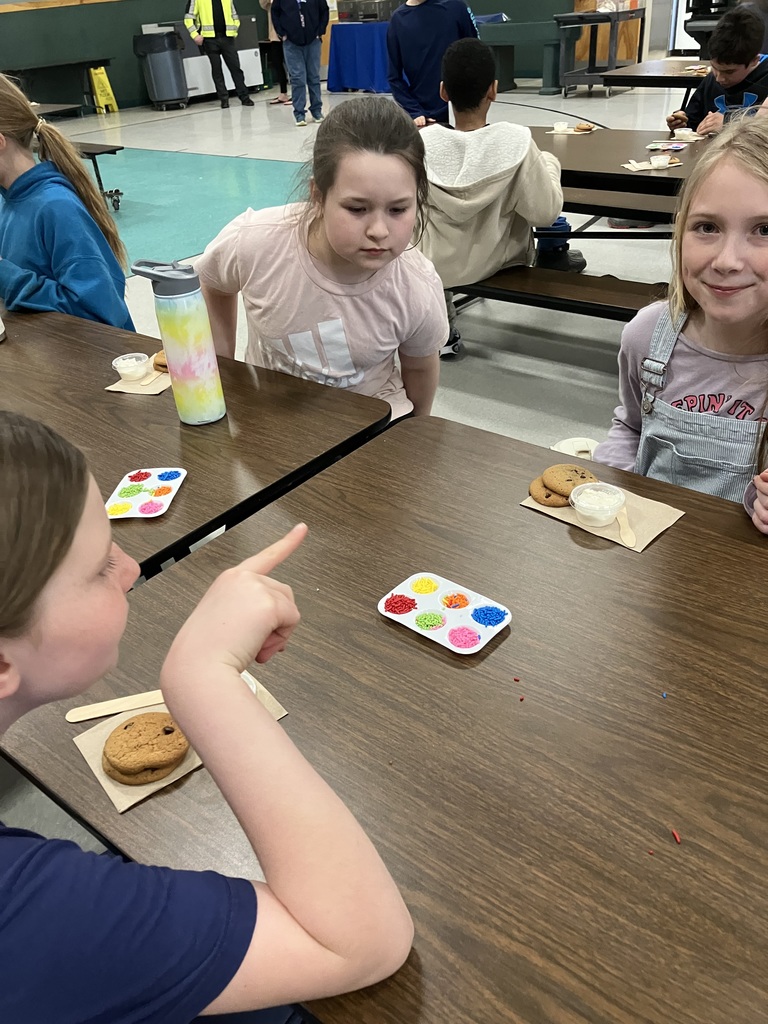  A close-up of three girls at a decorating station. One girl in a blue shirt points toward a palette of sprinkles in the center of the table. To the right, a girl in denim overalls smiles at the camera, while another girl in a light pink shirt looks on. Each child has their own set of cookies and frosting ready for decorating.